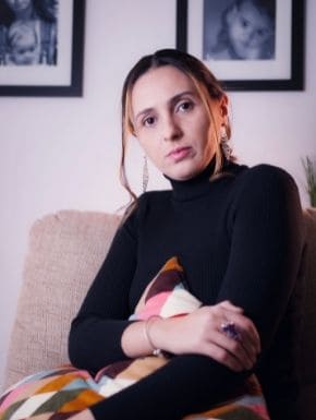Fotografia de Juliana Cascardo vestindo um suéter preto, sentada em um sofá e abraçando uma almofada colorida