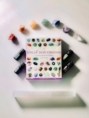 Fotografia do livro Bíblia dos Cristais, acima dela sete cristais com as sete cores do arco-íris, abaixo um cristal retangular e quatro florais, dois de cada lado